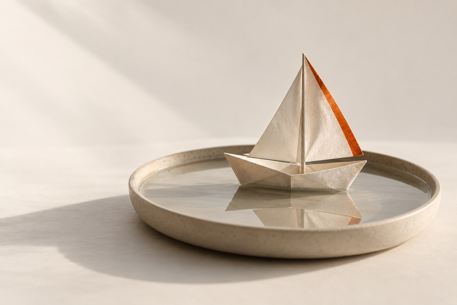 A paper sailboat with a burnt-orange sail edge floats in a shallow round tray of water.