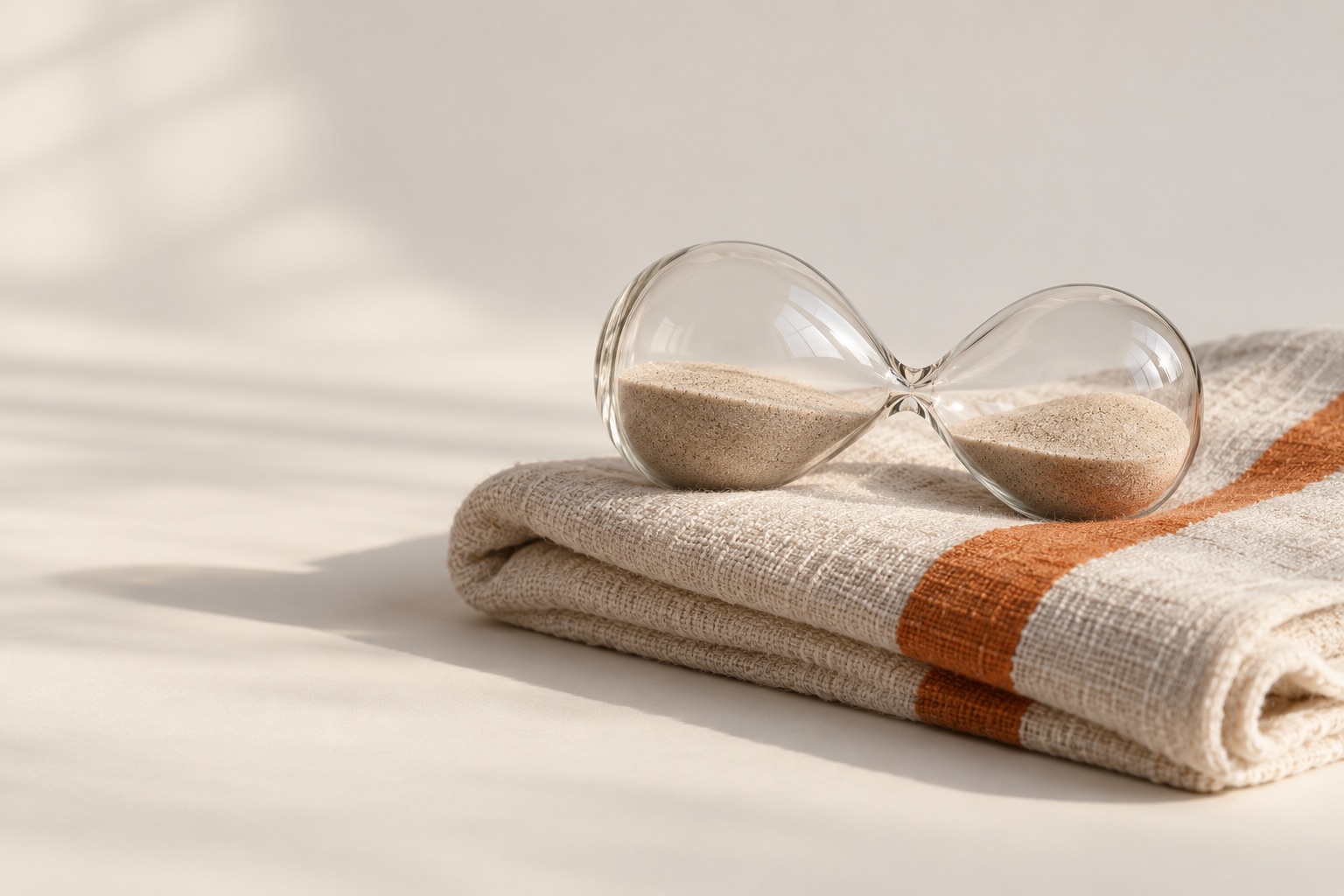 A glass hourglass lying on its side on a linen towel with a single orange stripe.