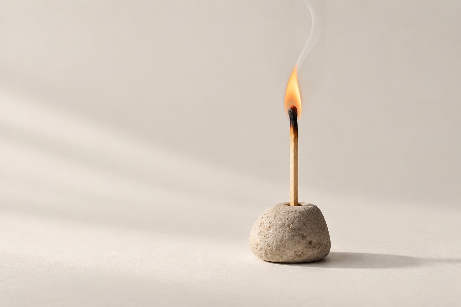 A burning wooden match stands upright in a rounded pale stone holder on a warm off-white surface.